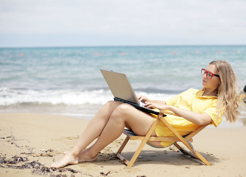 A girl sits in a deck chair at the beach with the sea behind her. She is wearing a bright yellow cover up. Learn how to stop glare on your screen - click the link below to read the blog post. MNK