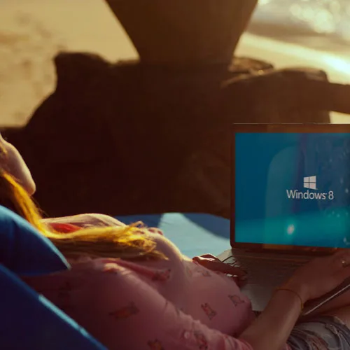A woman lays at the beach with her laptop on her lap, you can see glare on the screen from the sun. MNK
