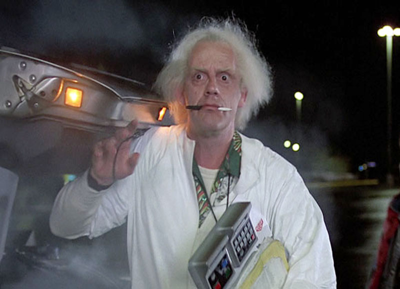 image of Doc Brown from the film Back To The Future with a pen in his mouth standing next to the Delorean car with it's door open